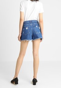Shorts de denim en azul medio con dobladillo deshilachado y detalles desgastados. Cuenta con dos bolsillos traseros y un diseño de cintura alta. Acompañados de tacones negros.