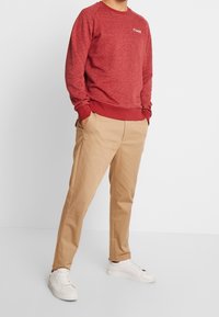 Sudadera roja texturizada con cuello redondo y puños acanalados, combinada con chinos beige y zapatillas blancas, con un corte relajado y bolsillos laterales.