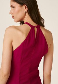 Mujer vista desde atrás que lleva un vestido sin mangas color magenta con cuello halter y cierre de botón en la nuca, pendientes de aro dorados visibles.