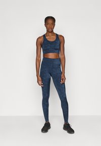 Compleu sport de culoare bleumarin, format dintr-un sutien sport ajustat și colanți cu talie înaltă, având un model texturat, asortați cu adidași negri.