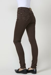 Jeans marrones para mujer con un diseño ajustado, que presentan un detalle de bolsillo en forma de corazón y una etiqueta en la cintura. Fabricados en denim con una textura suave.