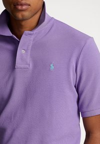Man i en lavendelfärgad polo med en delvis uppknäppt krage och en liten ljusblå broderad logotyp på vänster bröst.
