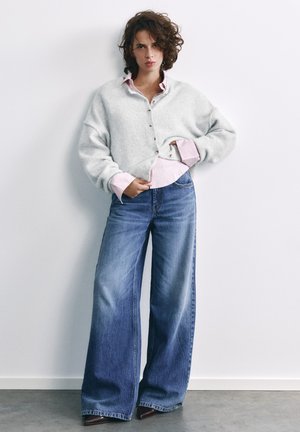 Femme aux cheveux bouclés portant un cardigan blanc boutonné par-dessus une chemise rose et un jean bleu à jambes larges, debout contre un mur blanc.