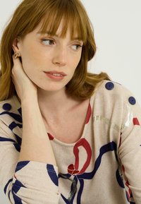 Top beige à manches longues avec des motifs abstraits colorés en bleu marine, rouge et accents métalliques ; présente des boutons bleu marine à l'épaule et une texture côtelée.