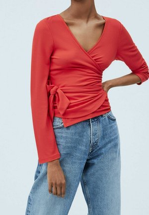 Kvinde iført en rød slå-om top med lange ærmer og en side sløjfe samt lyseblå højtaljede jeans, poserer med den ene hånd på hoften.