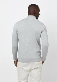 Grauer Strickpullover mit hohem Kragen und gerippten Bündchen, der eine glatte Textur aufweist. Über hellen Hosen getragen.