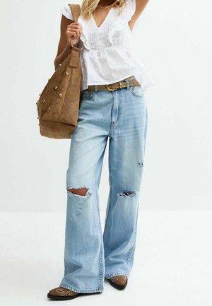 Woman wearing white ruffled blouse, light blue ripped wide-leg jeans, brown belt, brown boots, and carrying large brown studded shoulder bag.