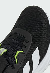 Czarne buty sportowe z teksturowaną siateczką na cholewce, zielonymi akcentami i białą gumową podeszwą. Posiada sznurowadła i wyściełany kołnierz dla wsparcia.