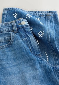 Blugi din denim albastru cu broderie florală albă, nasture metalic și buzunare standard, având o textură ușor decolorată.
