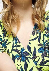 Persona bionda che indossa un vestito giallo con un motivo floreale blu e nero, che mostra il collo e la parte superiore del petto.