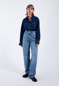 Dunkelblaue Jeanshemd mit zwei Fronttaschen, kombiniert mit hochgeschnittenen, hellblauen, weit geschnittenen Jeans und schwarzen Lederschuhen.