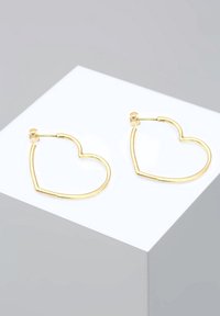 Paire de boucles d'oreilles fines en or en forme de cœurs, présentées sur un support géométrique blanc avec un fond gris.