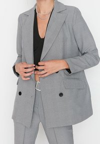 Šedý kostkovaný blazer s propracovaným střihem, který má klopový límec, dvojité zapínán�í na knoflíky a dvě přední kapsy pro dodatečný detail.