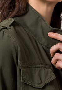 Oliven grøn jakke med en struktureret krave, skulderlapper og en klaplomme. Stoffet ser glat ud med en let tekstureret overflade.