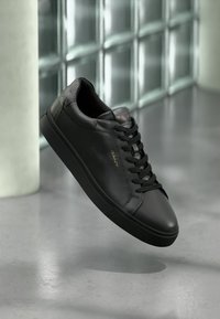 Adidași negri din piele cu șireturi negre și logo-ul "GANT" în aur, prezentați plutind deasupra unei pardoseli din beton lăcuit, aproape de un perete din blocuri de sticlă.