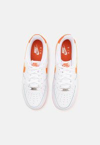 Bijele tenisice Nike Air Force 1 imaju narančaste akcentne boje i teksturirani dizajn, s perforiranim prednjim dijelom i ravnim vezicama.