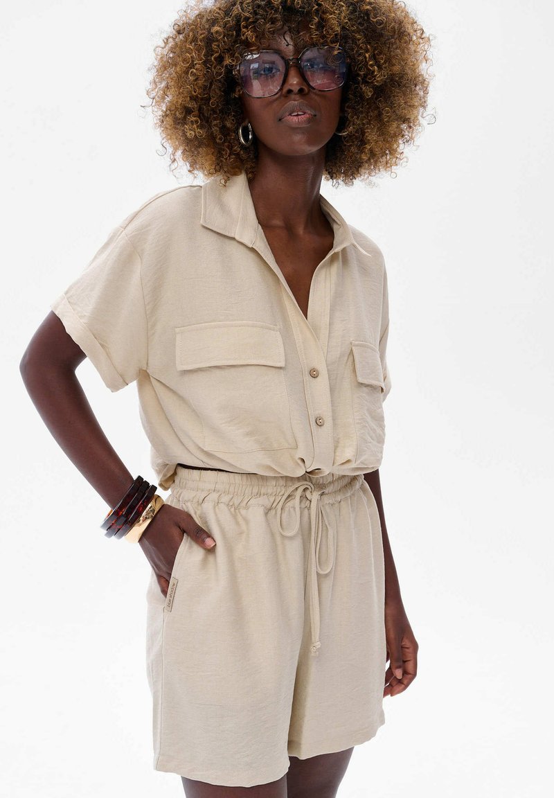 Beige two-piece outfit featuring a short-sleeve button-up top with pockets and drawstring shorts. Smooth fabric and relaxed fit.