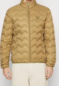 Hombre vistiendo una chaqueta acolchada color canela con cierre delantero y un pequeño logo de ave amarilla en el lado izquierdo del pecho, combinada con pantalones beige claros.