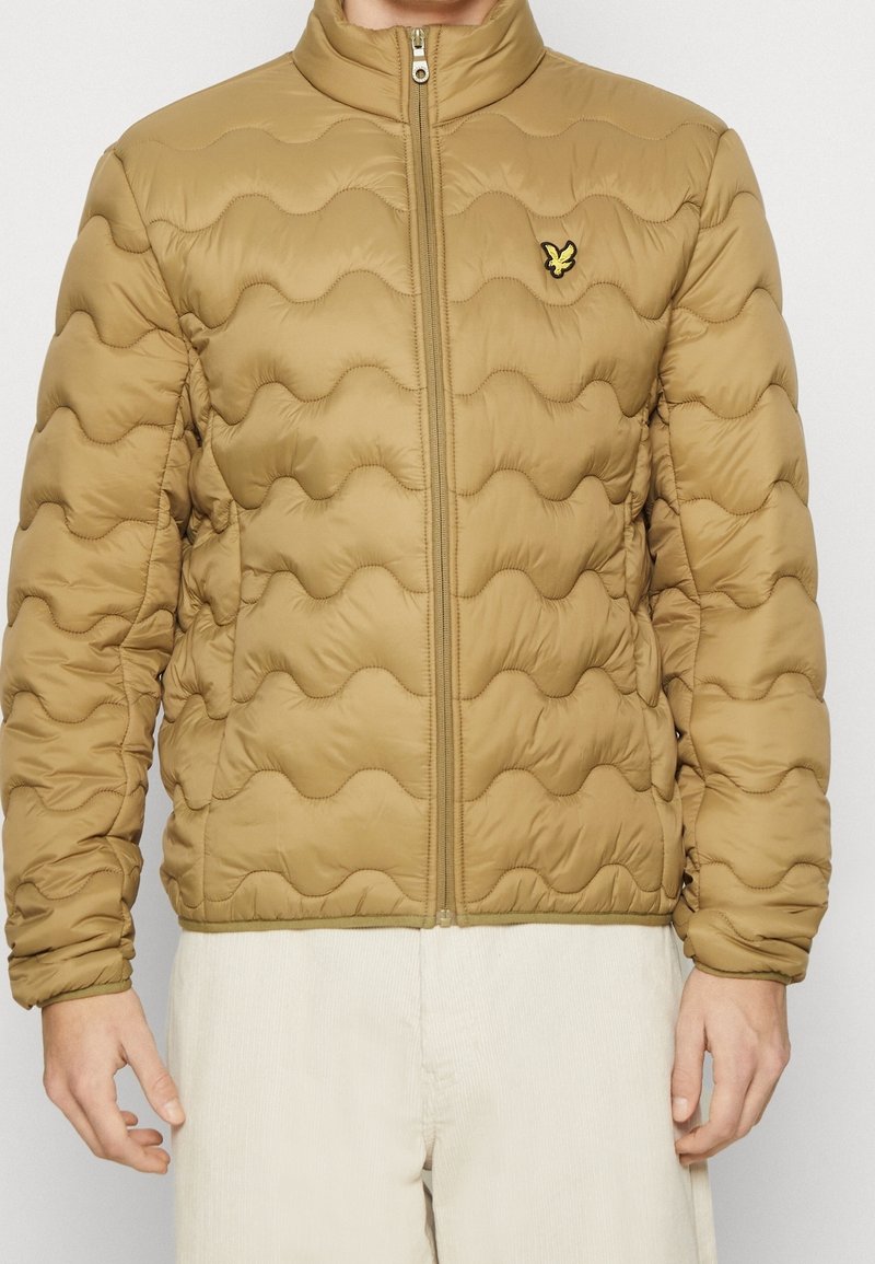 Hombre vistiendo una chaqueta acolchada color canela con cierre delantero y un pequeño logo de ave amarilla en el lado izquierdo del pecho, combinada con pantalones beige claros.