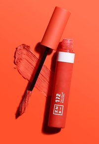 Korallfarget mascara i en glinsende tube med en matt hette. Har en hvit stripe med produktinformasjon. Follows med en børste og en prøve av produktet.