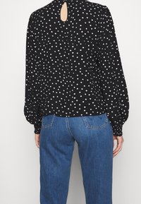 Svart blus med vita prickar och långa puffärmar, med nyckelhålsdetalj bak, ihopparad med ljusblå högmidjade jeans. Mjuk tygkänsla.