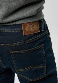 Vaqueros de mezclilla azul oscuro con un parche de cuero que presenta un diseño en relieve, costuras amarillas y dos bolsillos traseros con costuras curvas.