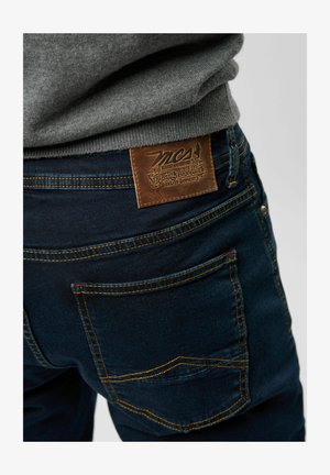 Dunkelblaue Denim-Jeans mit einem Lederpatch mit geprägtem Design, gelben Nähten und zwei hinteren Taschen mit geschwungenen Nähten.