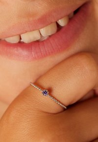 Silberner Ring mit einem perlengeschmückten Band, das einen zentralen lila Edelstein zeigt, platziert auf einer Hand in der Nähe eines lächelnden Mundes mit natürlichen Lippen.