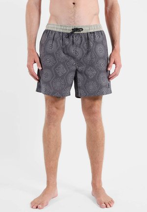 Homme portant un short de bain gris à motifs avec une ceinture élastique beige et un cordon noir, debout pieds nus sur fond blanc.