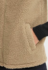 Veste en polaire beige avec un extérieur texturé et doux, présentant un poignet en tricot noir et une poche latérale. Fermeture éclair à l'avant.