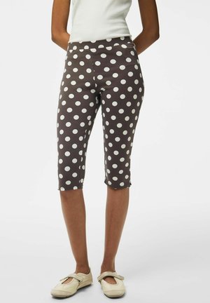 Femme portant des leggings marron mi-genoux à pois blancs et des chaussures plates beiges, debout les bras derrière le dos.