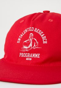Punane kork, mis on valmistatud tekstuurses kangas, esitleb valget graafilist disaini mehesse, kes kannab kaitseprille ja jälgib arvutit. Tekst ütleb "UNTALENTED RESEARCH PROGRAMME."