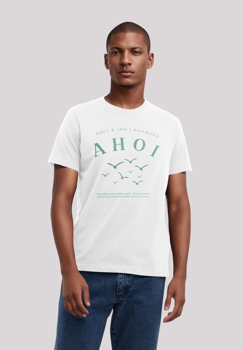 Weißes Baumwoll-T-Shirt mit "AHOI" in grün, grafischen Vögeln und Text darunter. Runder Hals und kurze Ärmel. Kombiniert mit blauen Jeans.