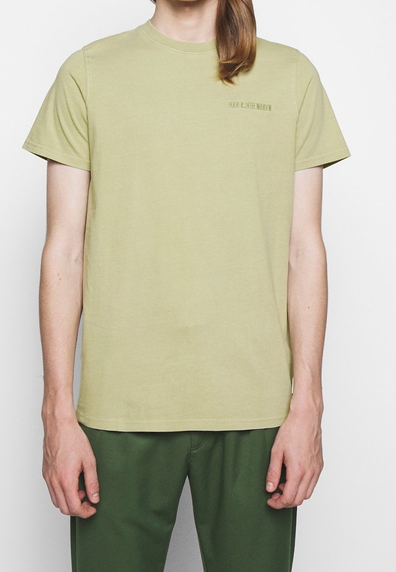 Person som bär en ljus olivgrön kortärmad T-shirt med liten broderad text på bröstet, i kombination med mörkgröna byxor.