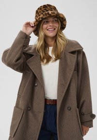 Smeđi vuneni kaput s širokim ovratnikom i duplim dugmadima, uparen s šeširom u uzorku leoparda i bijelom majicom.