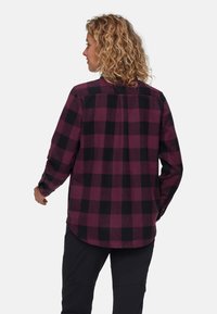 Camicia in flanella a scacchi viola e nera con maniche lunghe e polsini con bottoni; texture liscia e orlo curvo. Indossata da una persona vista da dietro.