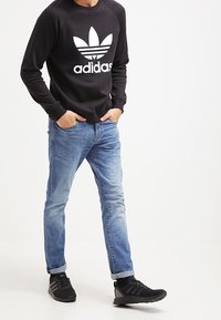 Svart sweatshirt med vitt Adidas-logotyp, ljusblå jeans och svarta joggingskor. Sweatshirten har rundad halsringning och ribbade ärmmuddar.