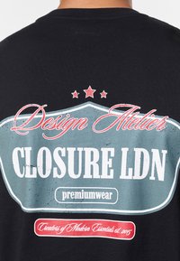 Svart t-shirt med en färgstark grafisk design på baksidan med text i vitt och rött, inramad av en teal-färgad märkesemblem med tre stjärnor ovanför.