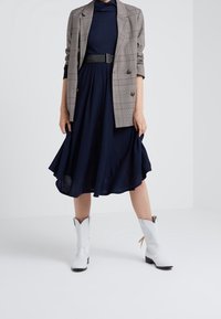 Grå rutiga kavaj, marinblå klänning med utställd kjol och vita cowboystövlar med svarta detaljer. Slätt tyg och klassiska designelement.