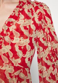Blouse rouge avec un motif paisley doré, tissu transparent, encolure à volants et détails en relief le long des manches.