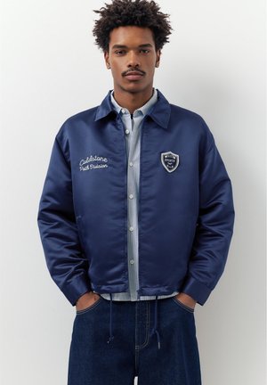 Ung mand med krøllet hår iført en marineblå satin sportjakke over en lyse stribet skjorte med knapper, hænderne i jeanslommerne, neutral baggrund.