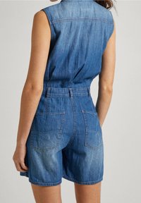 Bezramenný modrý deninový overal s zadními kapsami, který má na sobě osoba stojící na světlém jednobarevném pozadí.