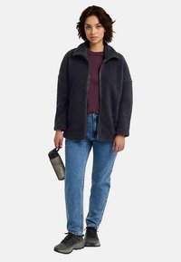 Donkergrijs fleecejack met een hoge kraag, gedragen over een bordeauxrode top, gecombineerd met een blauwe spijkerbroek en zwarte sneakers. Houdt een fles water vast.