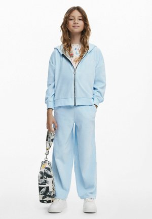 Hellblaues Sweatshirt und Hosen-Set aus weichem Material, mit einem Reißverschluss und gerippten Bündchen. Das Outfit wird mit einer gemusterten Tasche kombiniert.