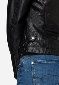 Svart läderjacka med en texturerad yta, siddragkedja och justerbar rem med metallknapp. Bärs över blå denimjeans.