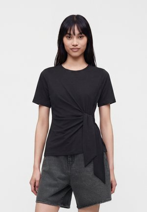 Jeune femme aux longs cheveux noirs portant un haut noir à manches courtes avec un nœud sur le côté et un short en denim gris foncé sur un fond uni.