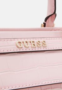 Sac à main en texture crocodile rose clair avec des lettres en métal doré formant le mot GUESS et une anse rose assortie.