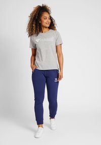Grijze T-shirt met korte mouwen en een wit logo, gecombineerd met marineblauwe joggers met zakken en witte details. Gekozen met witte sneakers.