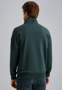 Dark green sweatshirt with a high collar, ribbed cuffs, and a patch pocket on the left arm, made from soft, textured fabric.