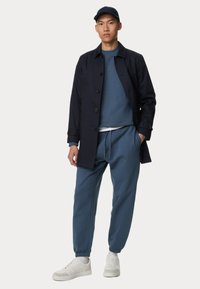 Cappotto trench blu navy, felpa blu, pantaloni jogger abbinati e sneaker bianche. L'outfit presenta una vestibilità moderna con un look casual e texture morbide.